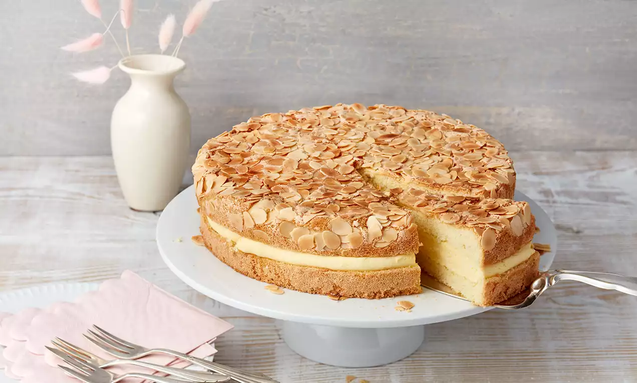 Okrugli kolač sa zlatnosmeđom koricom prekrivenom listićima badema, žutom puding kremom u sredini, na bijelom stalku za torte