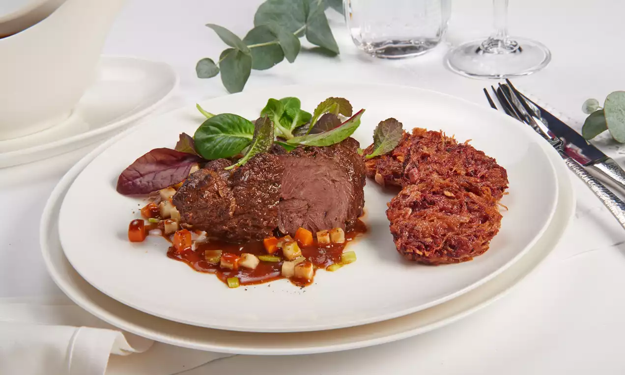 Geschmorte Kalbsbäckchen dunkelbraun und zart auf Gemüsewürfeln mit knusprigen Rösti und grünen Blättern auf weißem Teller angerichtet