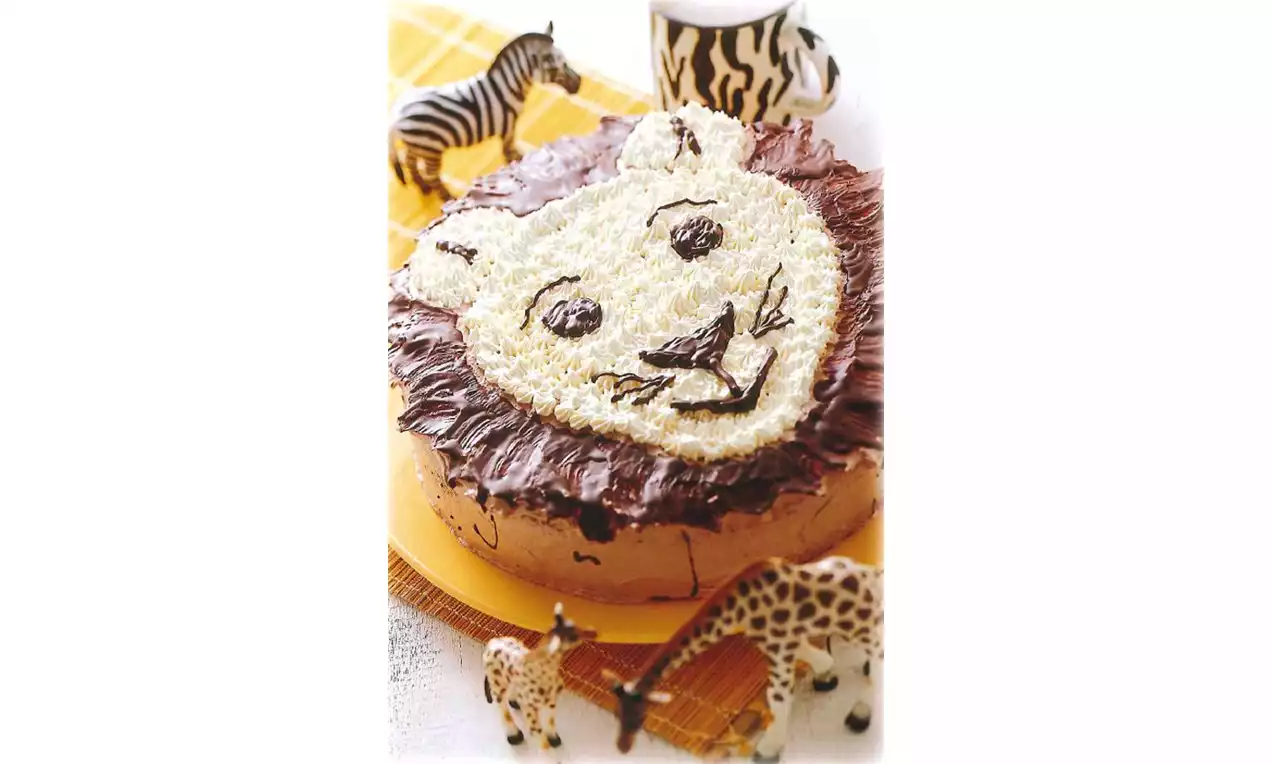 Gâteau rond recouvert de crème beige et chocolat formant un visage de lion, entouré de petites figurines d'animaux sur un tissu quadrillé