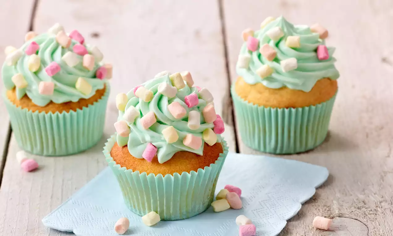 Vanillefarbene Muffins mit türkisgrünem Frosting, dekoriert mit bunten Mini-Marshmallows, in pastellblauen Papierförmchen auf Holzuntergrund