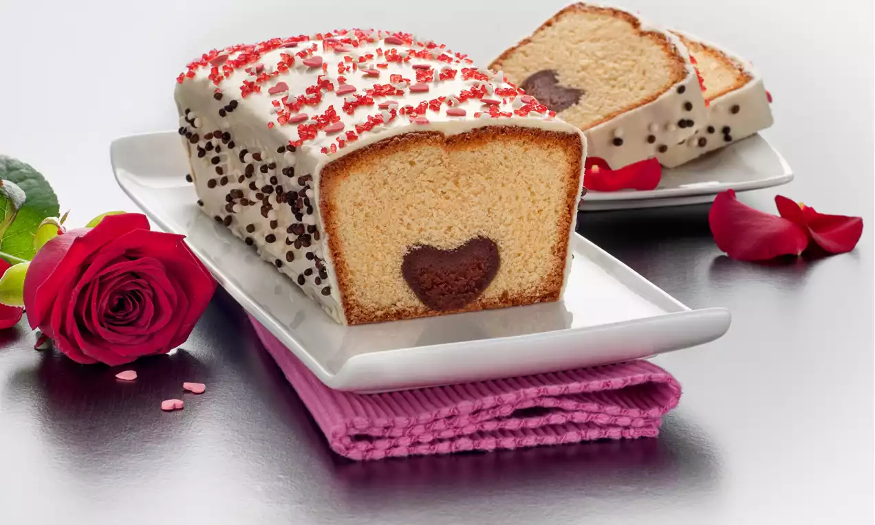 Chec auriu glazurat cu ciocolată albă, inimioară maro în secțiune, decorat cu inimioare roz și ciocolată, pe un platou alb