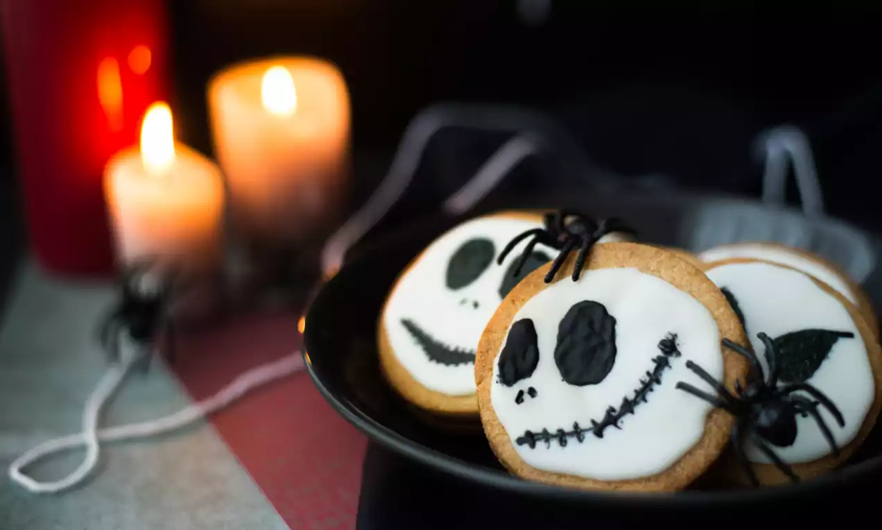 Ronde lichte koekjes met witte glazuurlaag, zwarte gezichten en decoraties, liggend op donker bord naast kaarsen en spinnen van touw