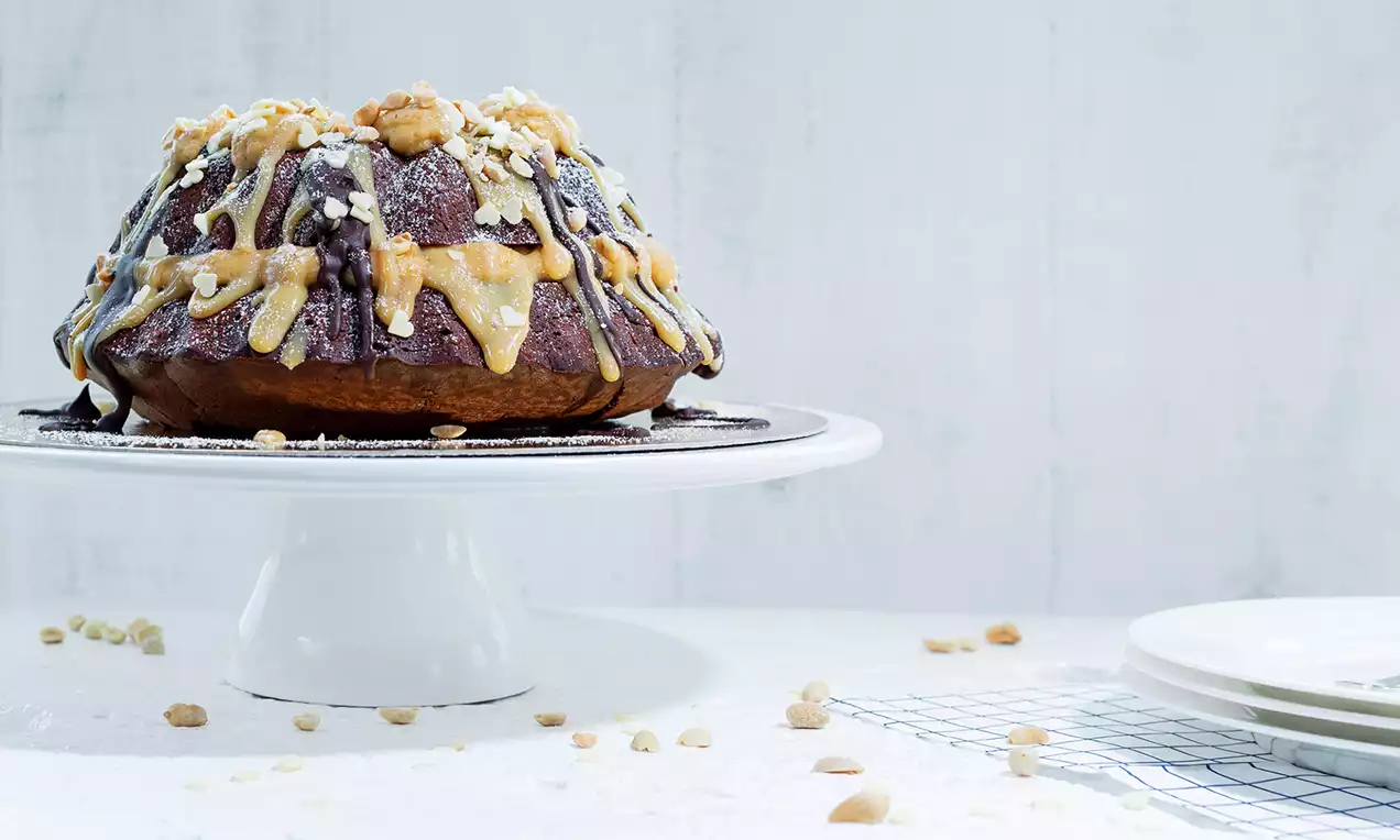 Chocolade Snickerstaart met glanzende donkere glazuur, lichte karamelstrepen, gehakte nootjes en een goudbruine rand op een wit taartplateau