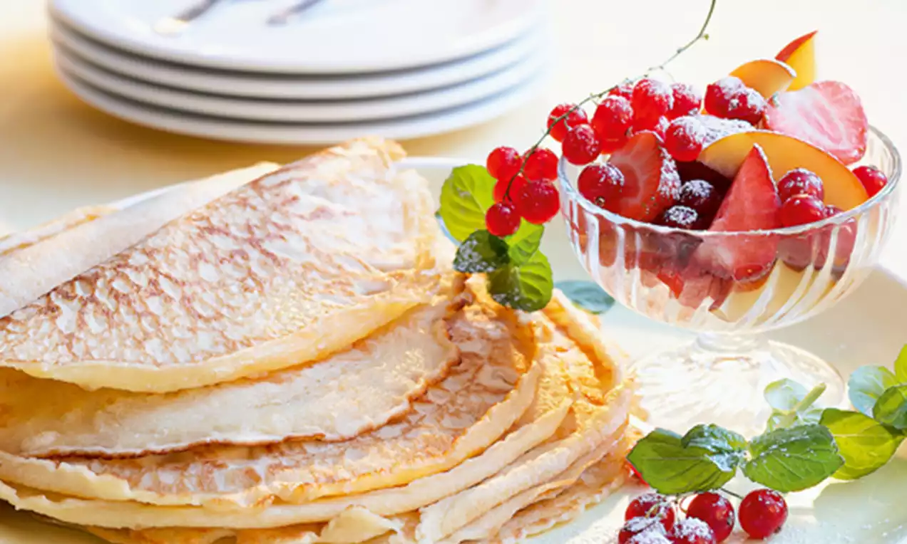 Crêpes fines et dorées empilées sur une assiette blanche, accompagnées de fruits rouges et jaunes frais dans une coupe transparente