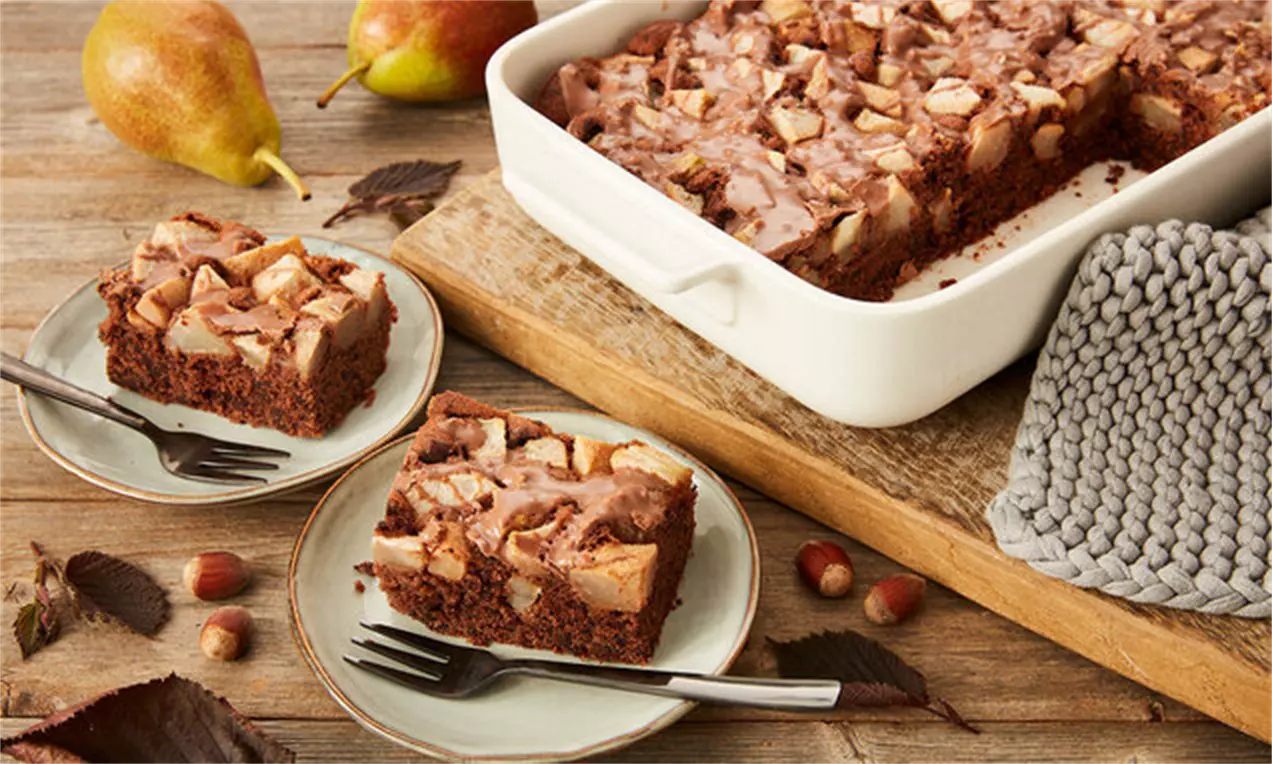 Кафяви квадратни парчета шоколадов сладкиш с меки парченца круши, подредени в бяла тава и малки десертни чинии на дървена маса