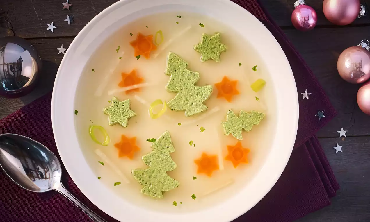 Klare Brühe mit grünen Broteinlagen in Form von Tannenbäumen, orangefarbenen Möhrensternen und feinen Lauchringen auf weißem Teller