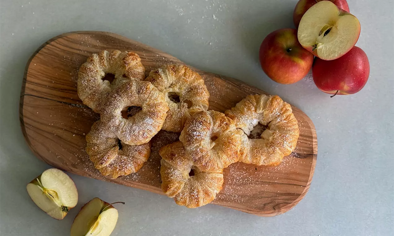 Goldbraune Apfel-Blätterteig-Ringe mit gezuckerter Oberfläche, angerichtet auf einem länglichen Holzbrett neben roten Äpfeln und Apfelspalten