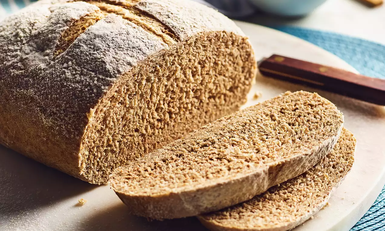 Round wholemeal loaf with a flour-dusted crust, golden brown interior and two slices cut, placed on a light plate with a textured blue mat