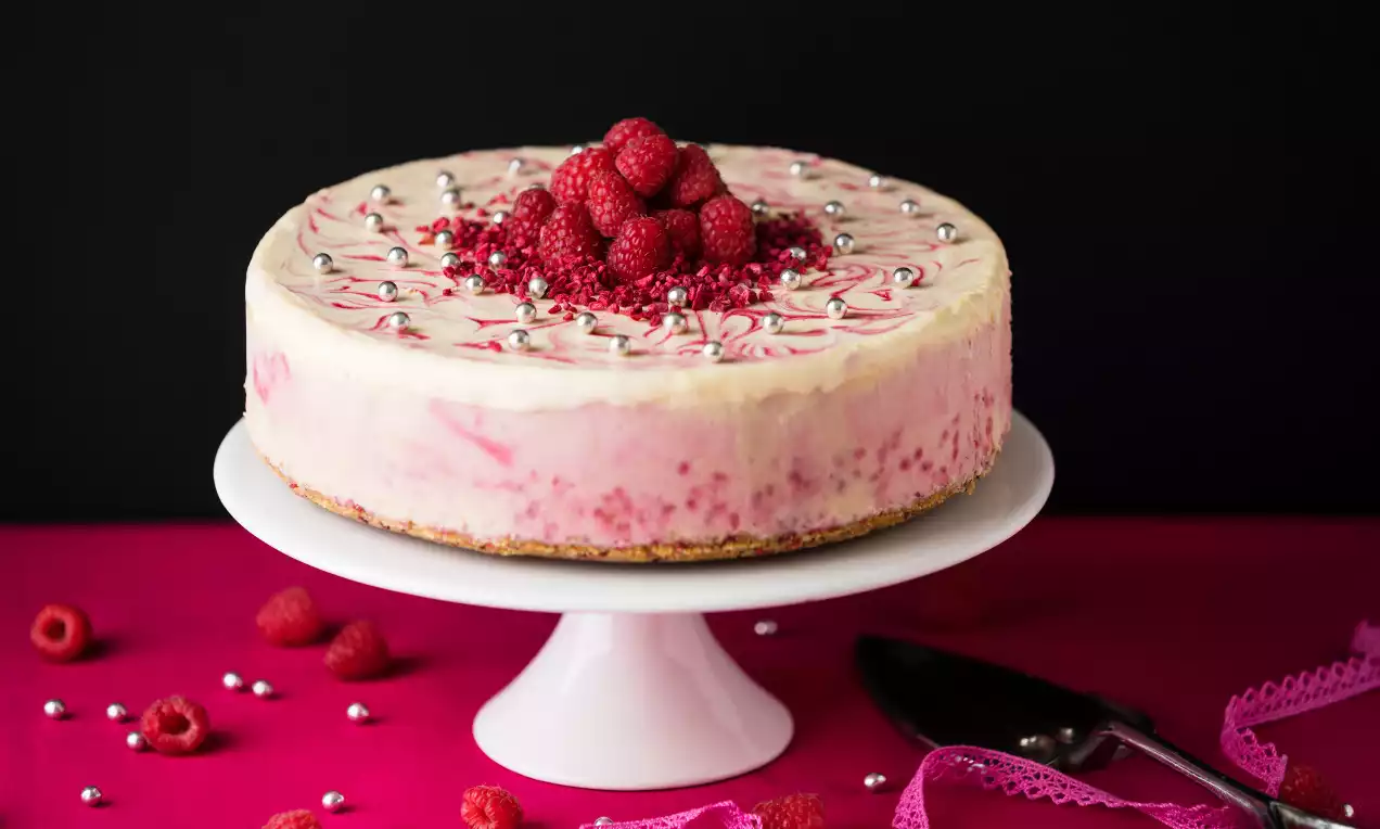 Rund tårta med rosa-marmorerad fyllning på en vit kakbotten, toppad med hallon, strössel och silverkulor på ett vitt tårtfat