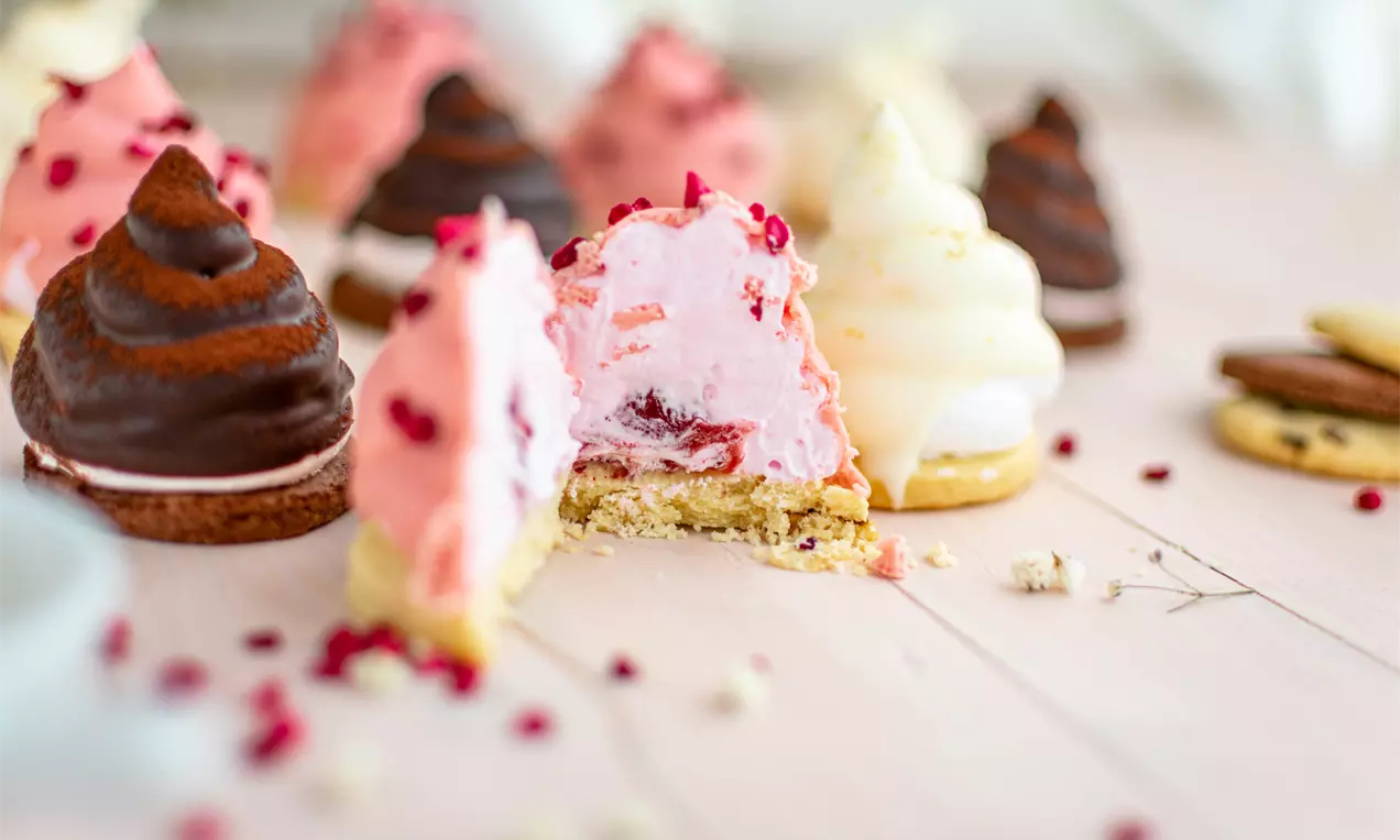 Чаени сладки със заоблени върхове в розово, кремаво и шоколадово кафяво, поставени върху светла дървена повърхност, с натрошени бисквити и червени плодчета