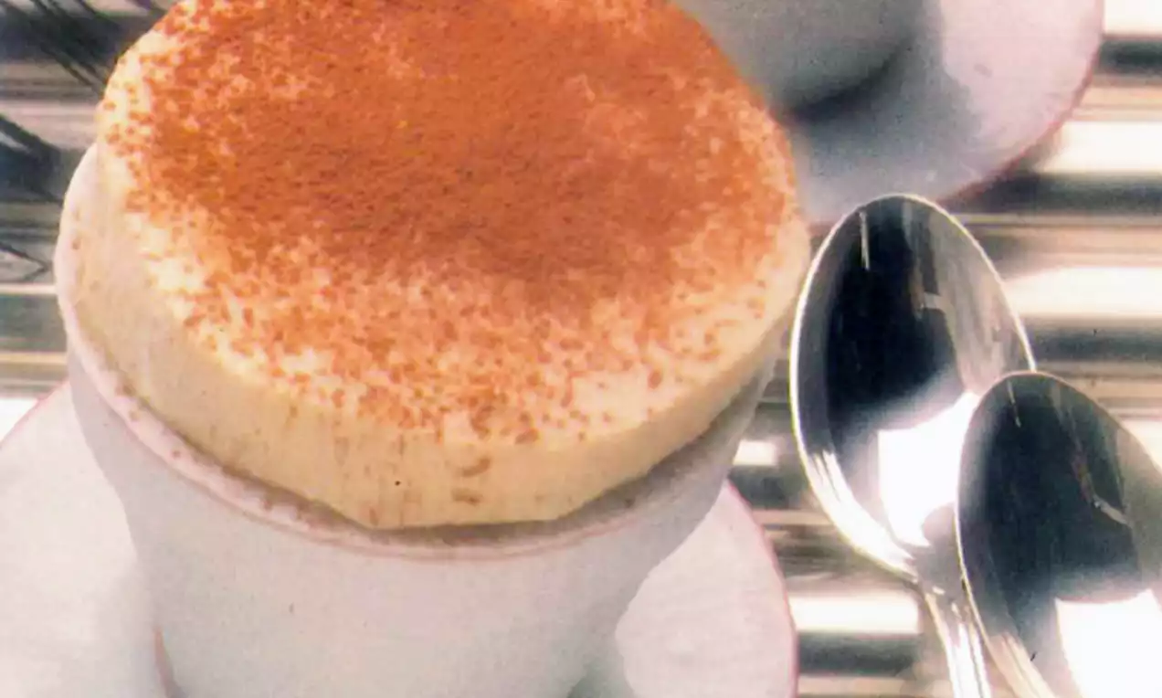 Bananenparfait met luchtige, crèmegele bovenlaag en fijnstrooisel van cacaopoeder in een witte kop op een schoteltje met twee lepels