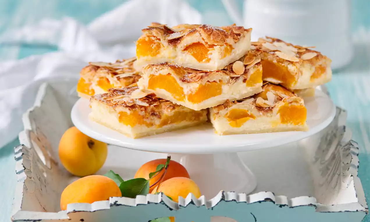 Čtverce světlého těsta s meruňkami a plátky mandlí vrstvené na bílém talíři, obklopené čerstvými meruňkami na světle modrém podkladu