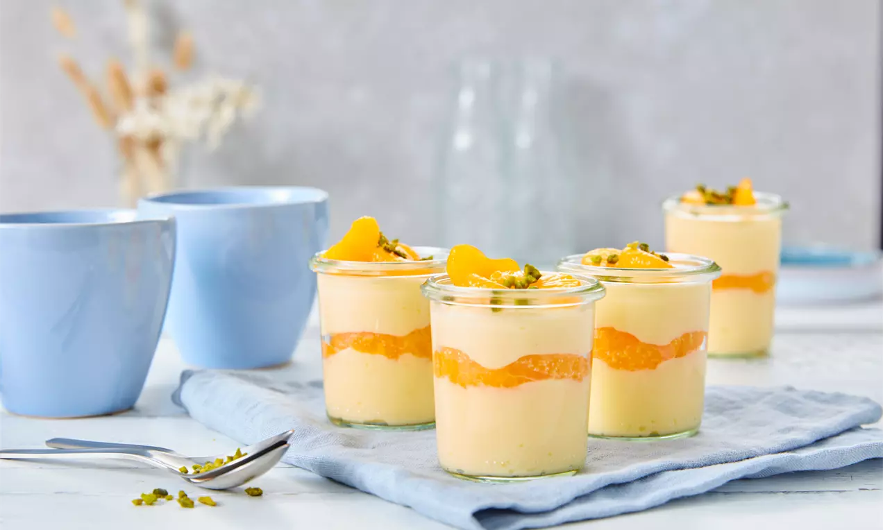 Heller Mandarinen-Cremepudding in transparenten Gläsern, geschichtet mit Orange-Streifen und Pistazien, auf blauem Tuch mit Tassen im Hintergrund