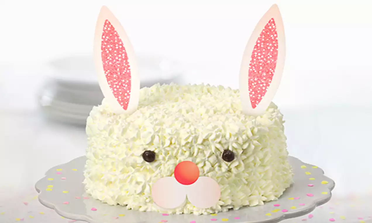 Tarta blanca decorada con crema rizada simulando un conejito, orejas rosas de papel, ojos oscuros y nariz redonda sobre un plato con confeti