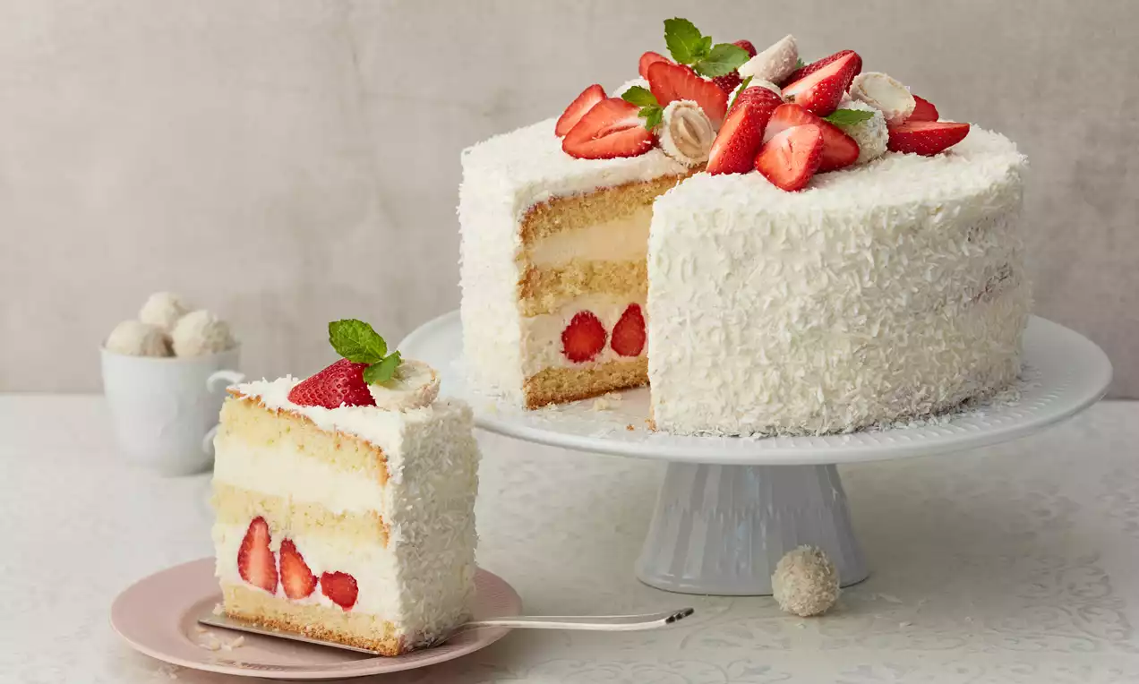 Bijela torta obložena kokosom, s dva sloja biskvita i kremom, ukrašena jagodama, Raffaello kuglicama i listićima mente na bijelom stalku