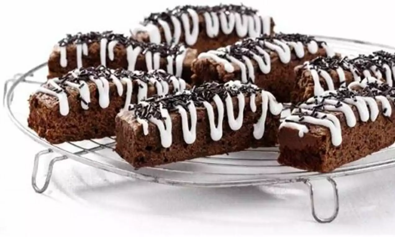 Negrese dreptunghiulare maro închis, pufoase, decorate cu glazură albă în zig-zag și fulgi de ciocolată, așezate pe un grătar metalic argintiu