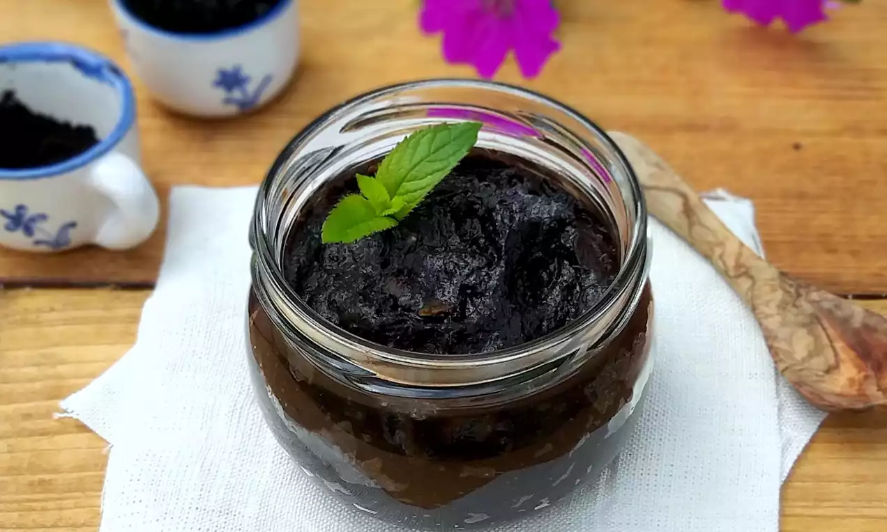 Tmavě hnědá švestková pomazánka s hustou texturou ve sklenici, ozdobená lístkem máty, na bílém ubrousku s dřevěnou lžící vedle