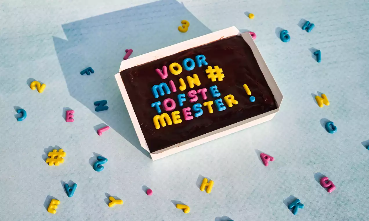 Donkerbruine brownie in een witte doos, versierd met kleurrijke lettertjes van fondant, op een lichtblauwe ondergrond bezaaid met losse letters