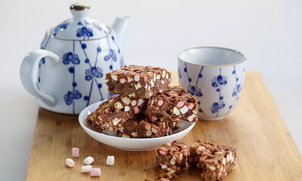 Carrés de gâteau au chocolat marbré de morceaux de marshmallows blancs et roses, empilés sur une assiette blanche avec un service à thé bleu et blanc en arrière-plan