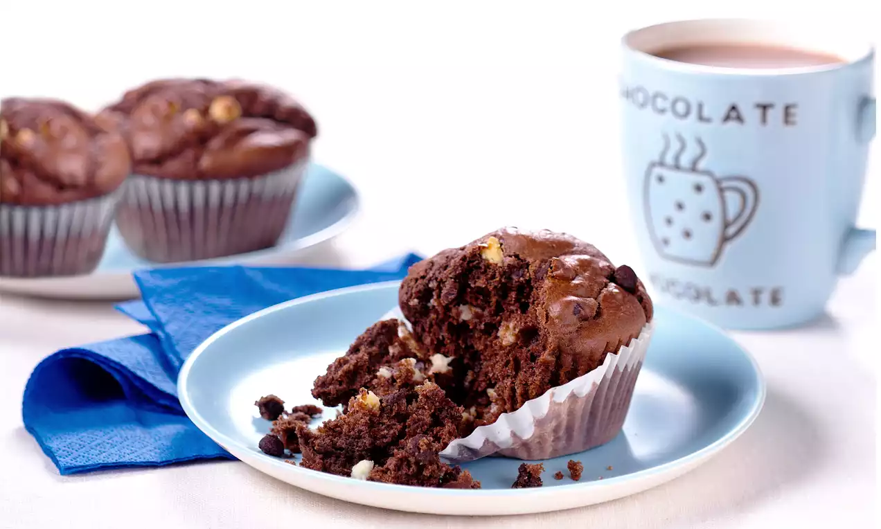 Aufgebrochener schokoladenbrauner Muffin mit cremigen weißen Schokostückchen auf hellblauem Teller neben Tasse und weiteren Muffins
