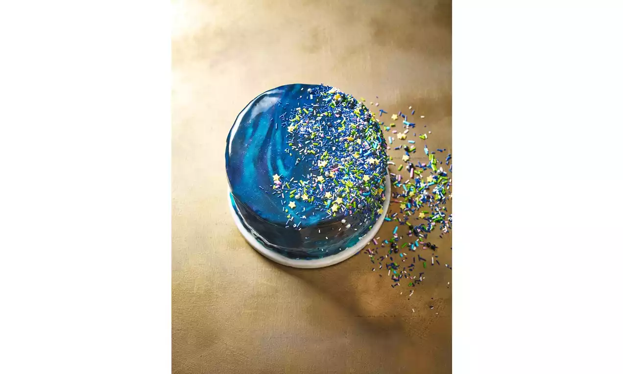 A round cake with a glossy blue mirror glaze swirled with shades of navy and turquoise, topped with scattered colorful sprinkles on gold