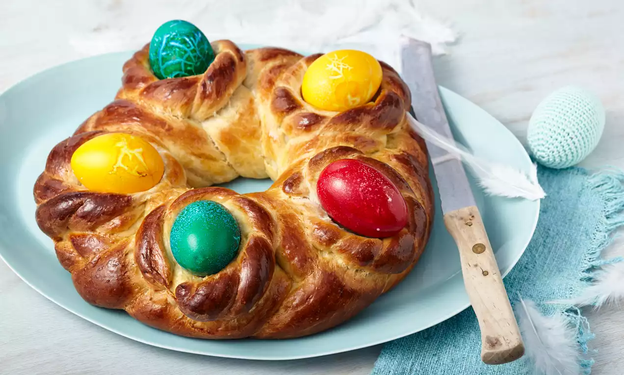 Couronne briochée dorée et brillante ornée de quatre œufs colorés rouge, jaune, bleu et vert sur une assiette bleu pastel