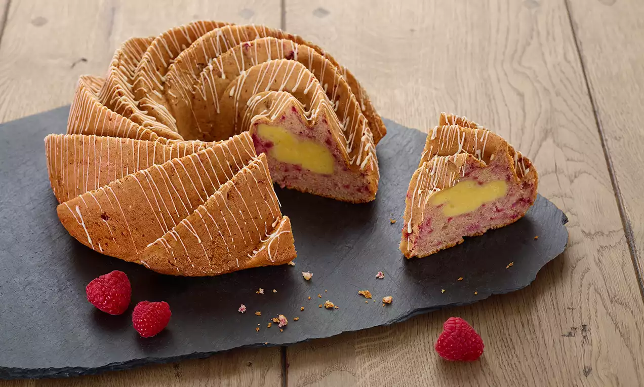 Gugelhupf doré aux motifs ondulés, garni de crème jaune vive et morceaux de framboises, posé sur une plaque sombre avec framboises fraîches
