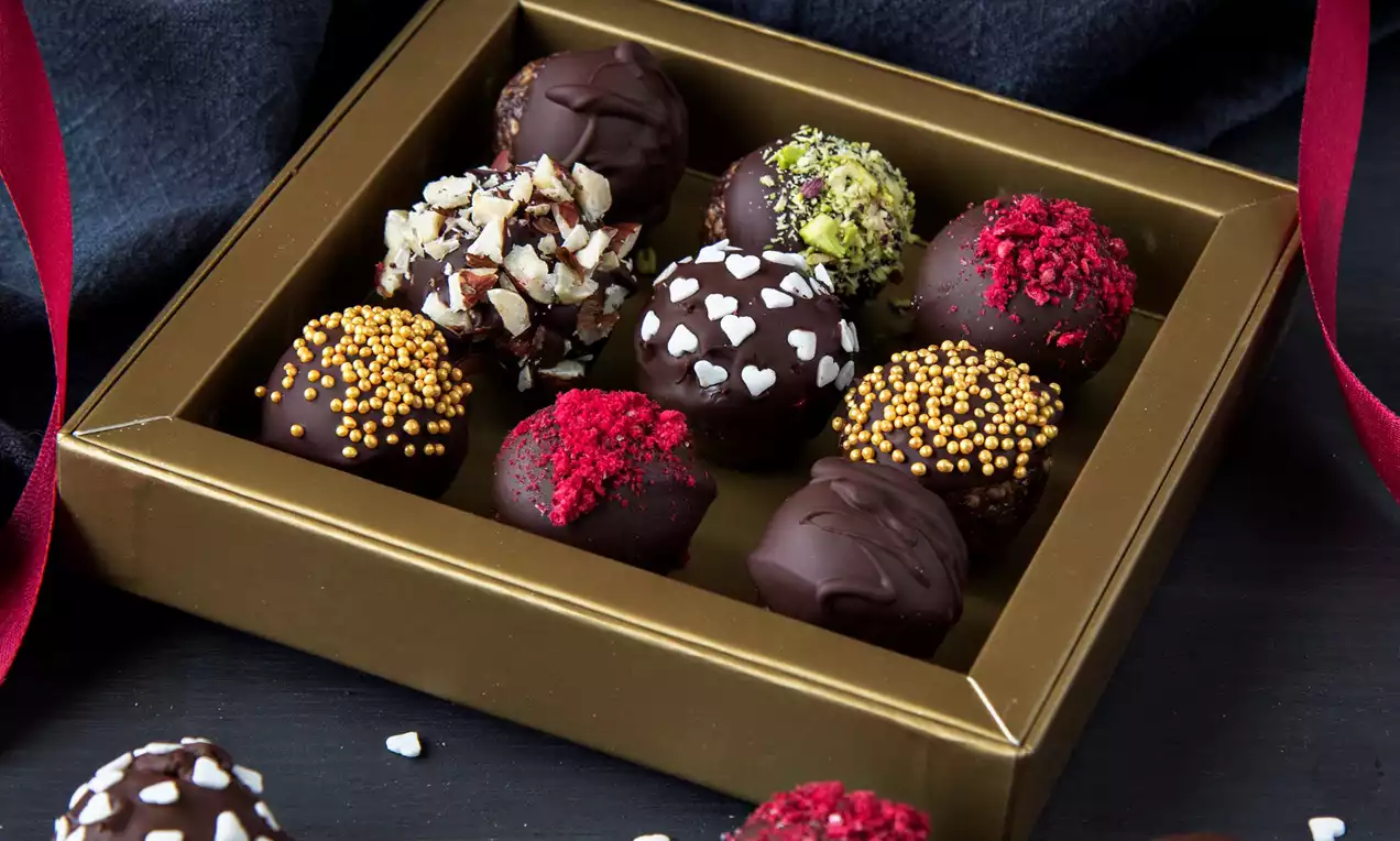 Čokoládové pralinky v tmavě hnědé zlato lemované krabičce, posypané různobarevnými ozdobami, okolní červené stuhy na černém pozadí