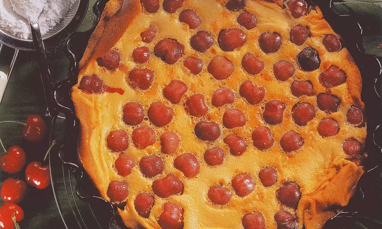 Clafoutis doré aux cerises rouges brillantes enfoncées dans une pâte moelleuse, présenté dans un moule noir sur une nappe verte