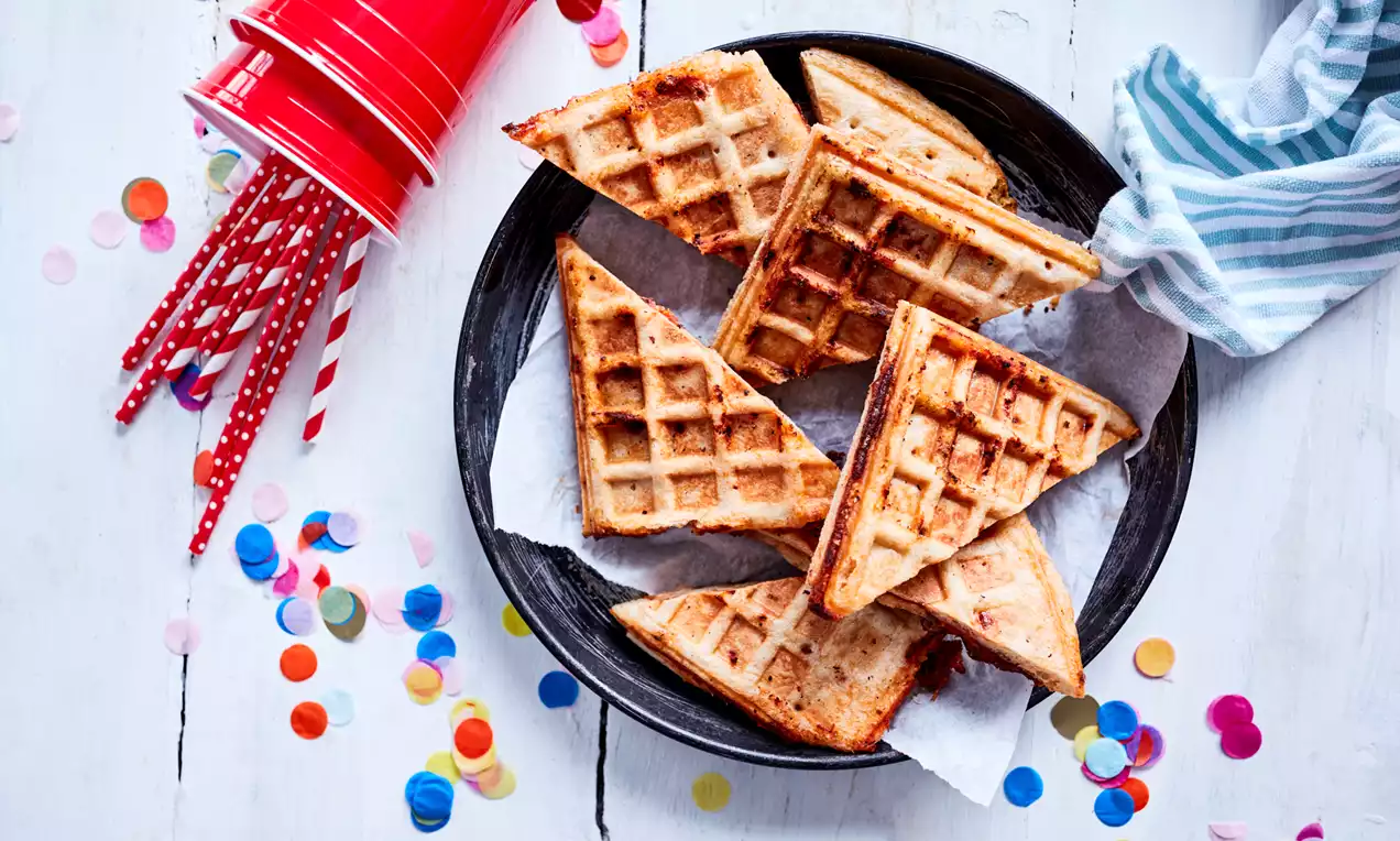 Goldbraune, dreieckige Pizza-Waffeln mit knuspriger Textur auf weißem Backpapier in schwarzem Teller, umgeben von bunten Konfetti