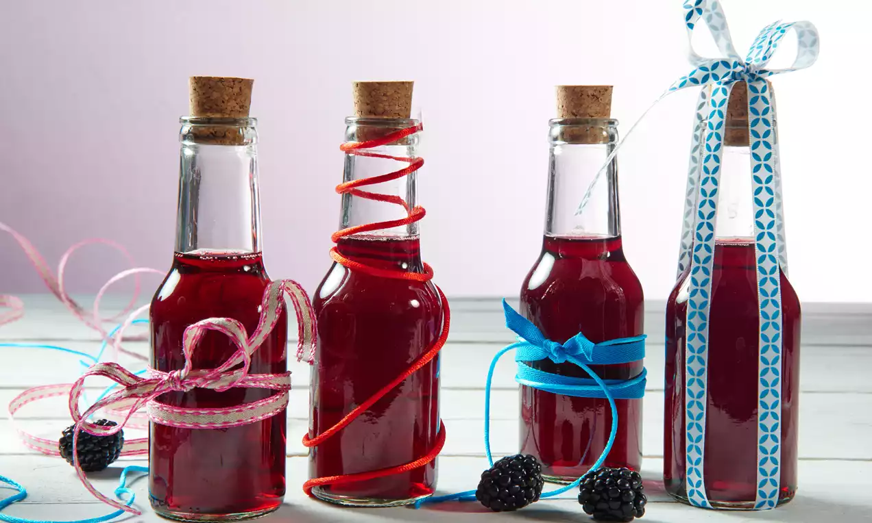 Vier schlanke Glasflaschen mit tiefrotem Brombeersirup, verziert mit bunten Schleifen, Korkverschlüssen und frischen Brombeeren als Dekoration