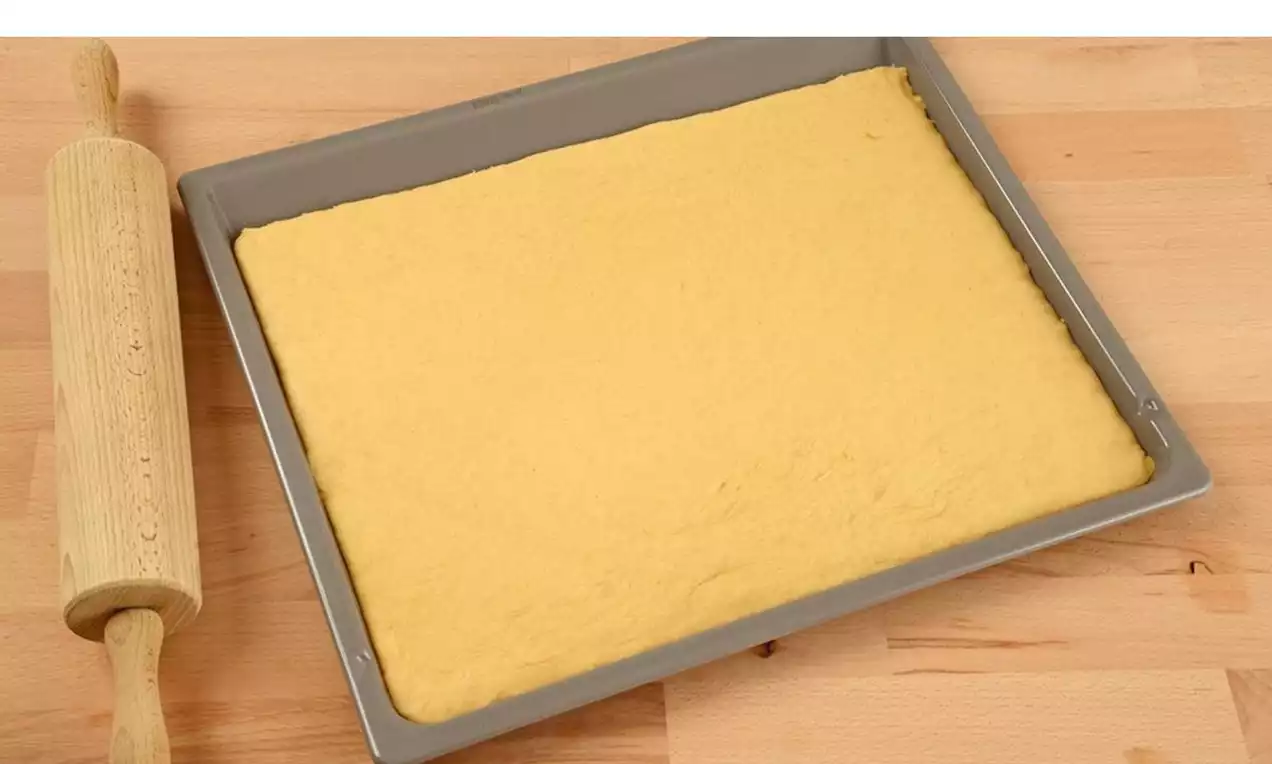 Goudgeel, glad uitgerold biscuitdeeg in een rechthoekige bakvorm met een houten deegroller op een licht houten werkblad