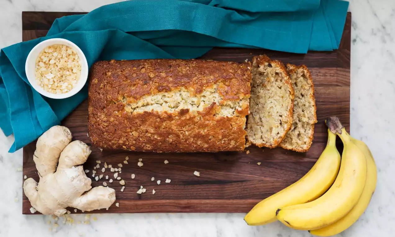 Goudbruin bananenbrood met een gebarsten korst, enkele plakken gesneden, op een houten plank naast verse gember, bananen en kandijstukjes