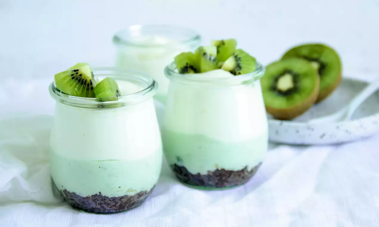 Slojevi svijetlozelene kreme i bijelog moussea u staklenki s tamnom podlogom, ukrašeni svježim kriškama kivija na bijeloj podlozi