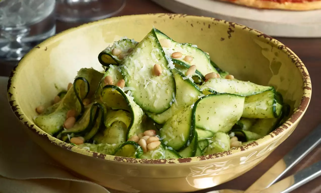Dunne, groene linten van courgette met een laagje pesto, gegarneerd met lichtbruine pijnboompitten in een beige kom met rustieke rand