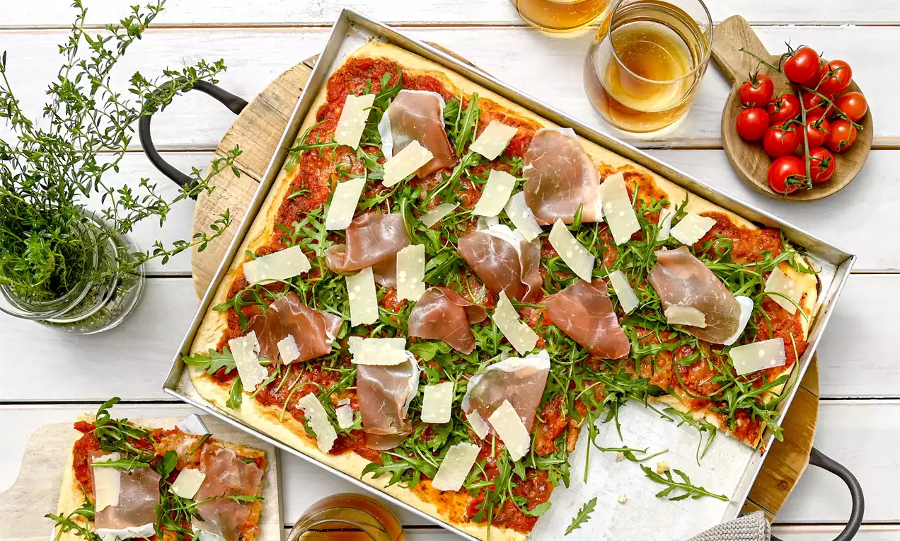 Pravougaona pizza na metalnom plehu s crvenim paradajz sosom, svijetlozelenom rikulom, tankim listovima pršute i kockicama sira