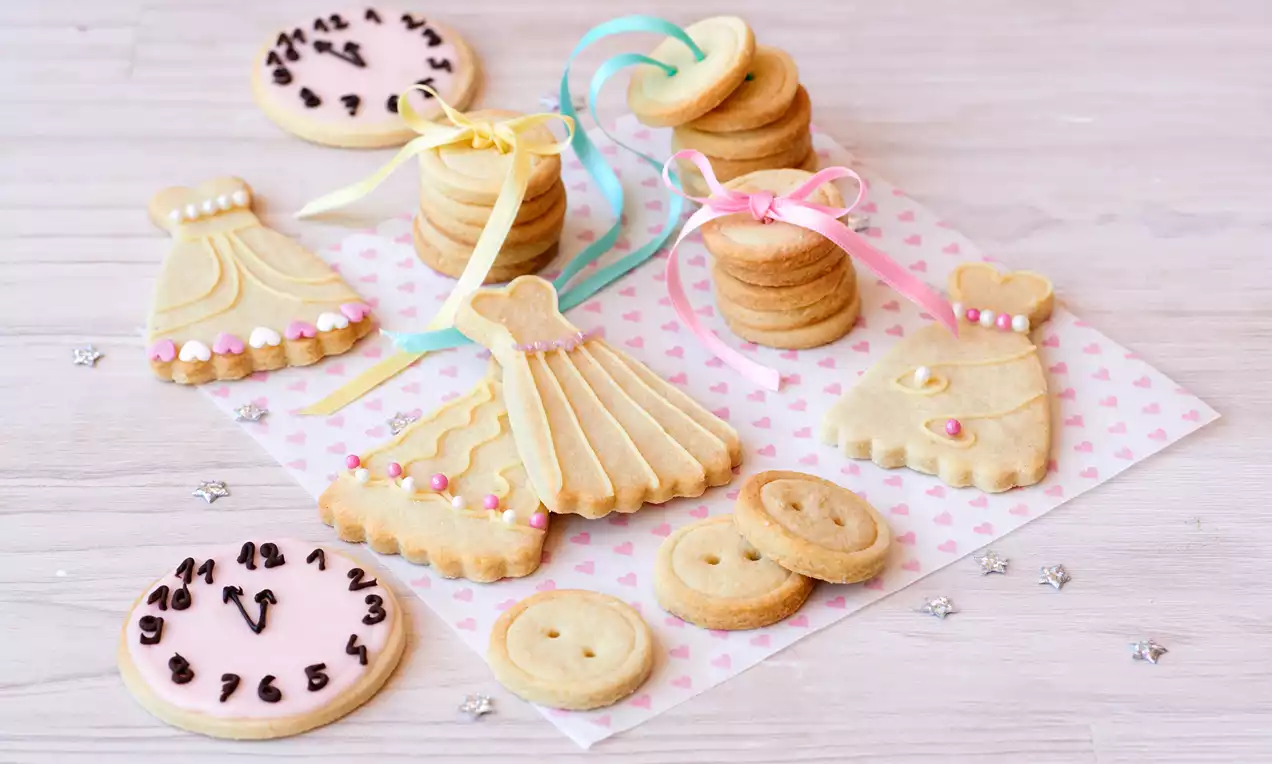 Galletas doradas en forma de vestidos decoradas con glaseado y perlitas, junto a otras redondas apiladas sobre papel de corazones rosas
