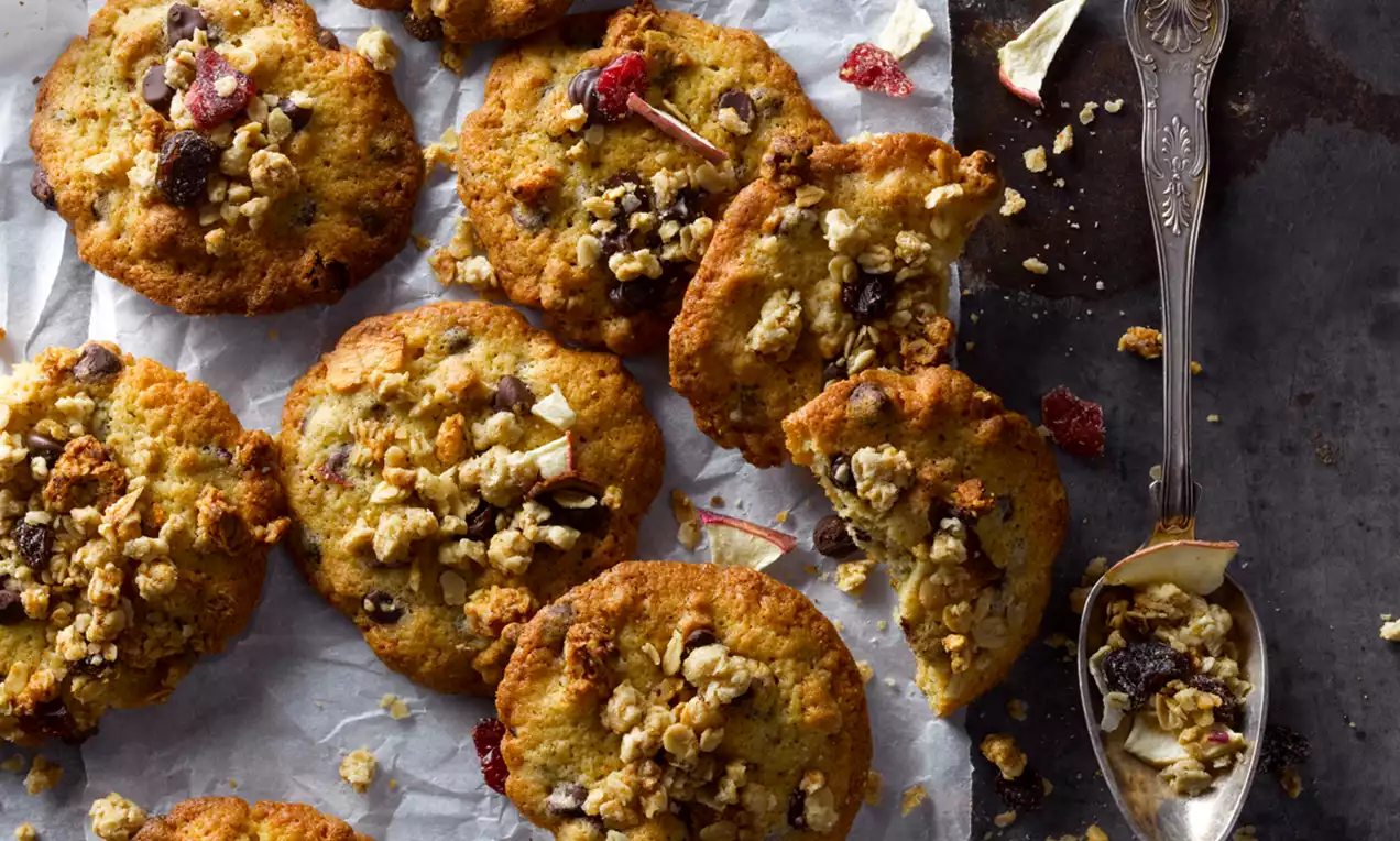 Rustiikkiset granolakeksit kullanruskeina, rapeareunaisina ja ruskeilla sekä punaisilla sävyillä koristeltuina leivinpaperilla ja metallilusikan kanssa