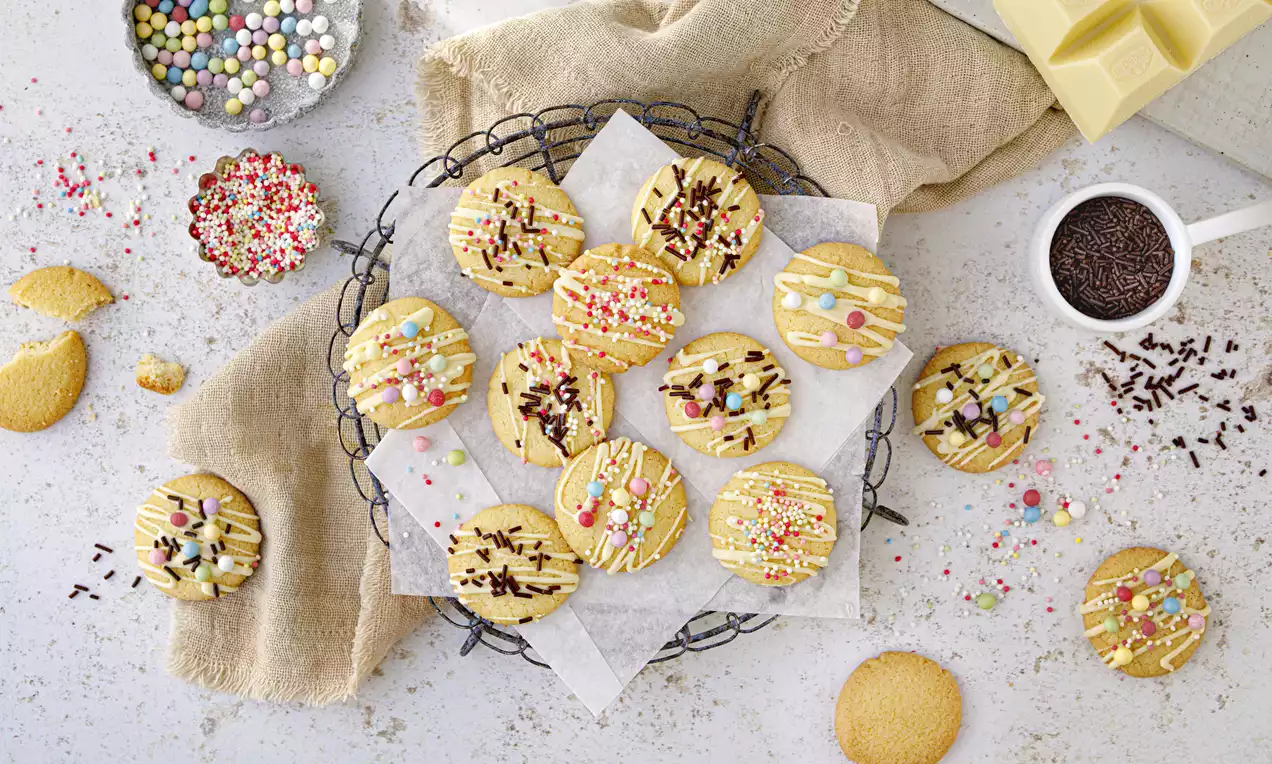 Hellgoldene Cookies mit bunter Verzierung aus Schokostreuseln, Zuckerperlen und Glasur auf weißem Backpapier auf einem Drahtgitter