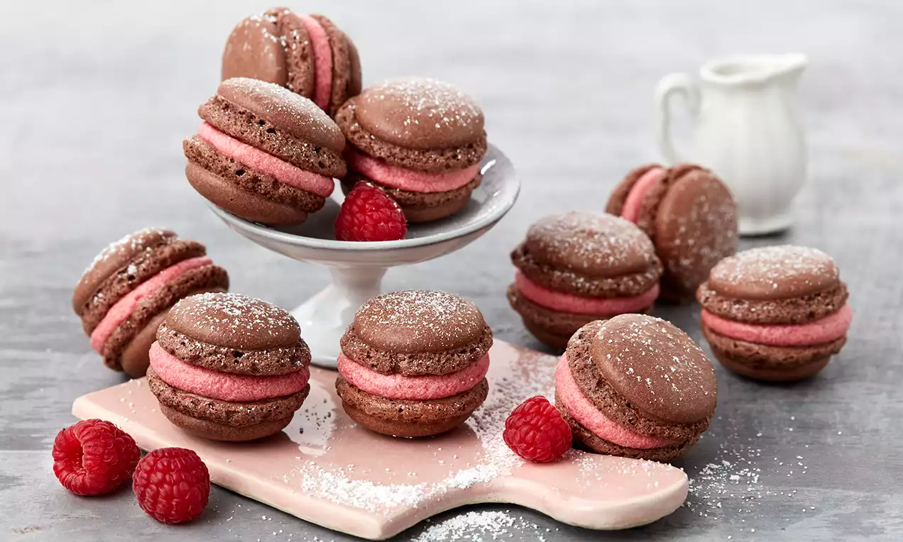 Čokoladni makaroni s glatkim malina kremom između dvije hrskave smeđe ljuskice, posluženi s malinama i posuti bijelim šećerom u prahu