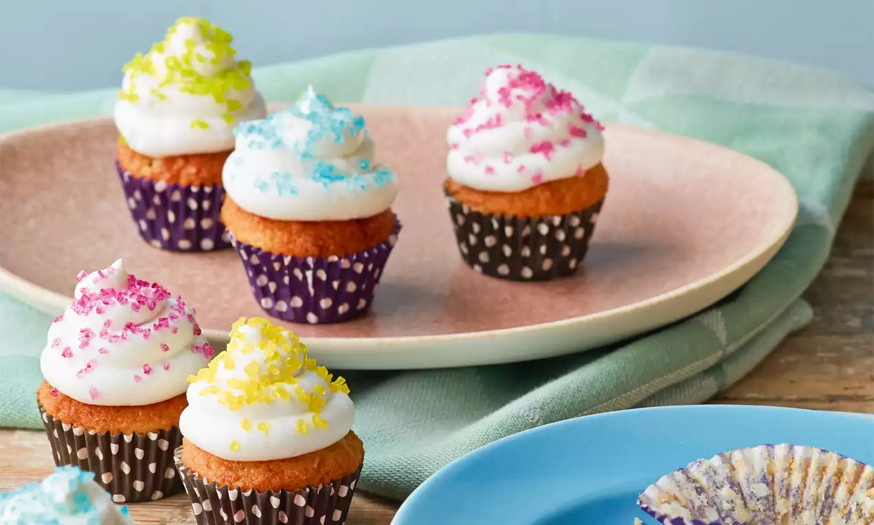 Mini-Pina-Colada-Cupcakes mit goldbraunem Teig, weißer Sahnehaube und buntem Zuckerdekor in gepunkteten Förmchen auf einem rosafarbenen Teller