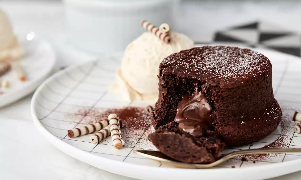 Dunkler, kreisförmiger Lava Cake mit glänzendem flüssigem Kern, bestäubt mit Puderzucker, auf einem Teller mit Eis und geschwungenen Dekorrollen