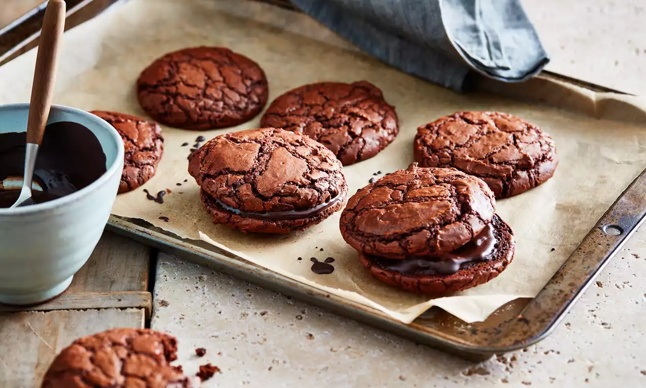 Cookies au chocolat brun foncé, aux bords légèrement craquelés, garnis de ganache, disposés sur une plaque recouverte de papier cuisson