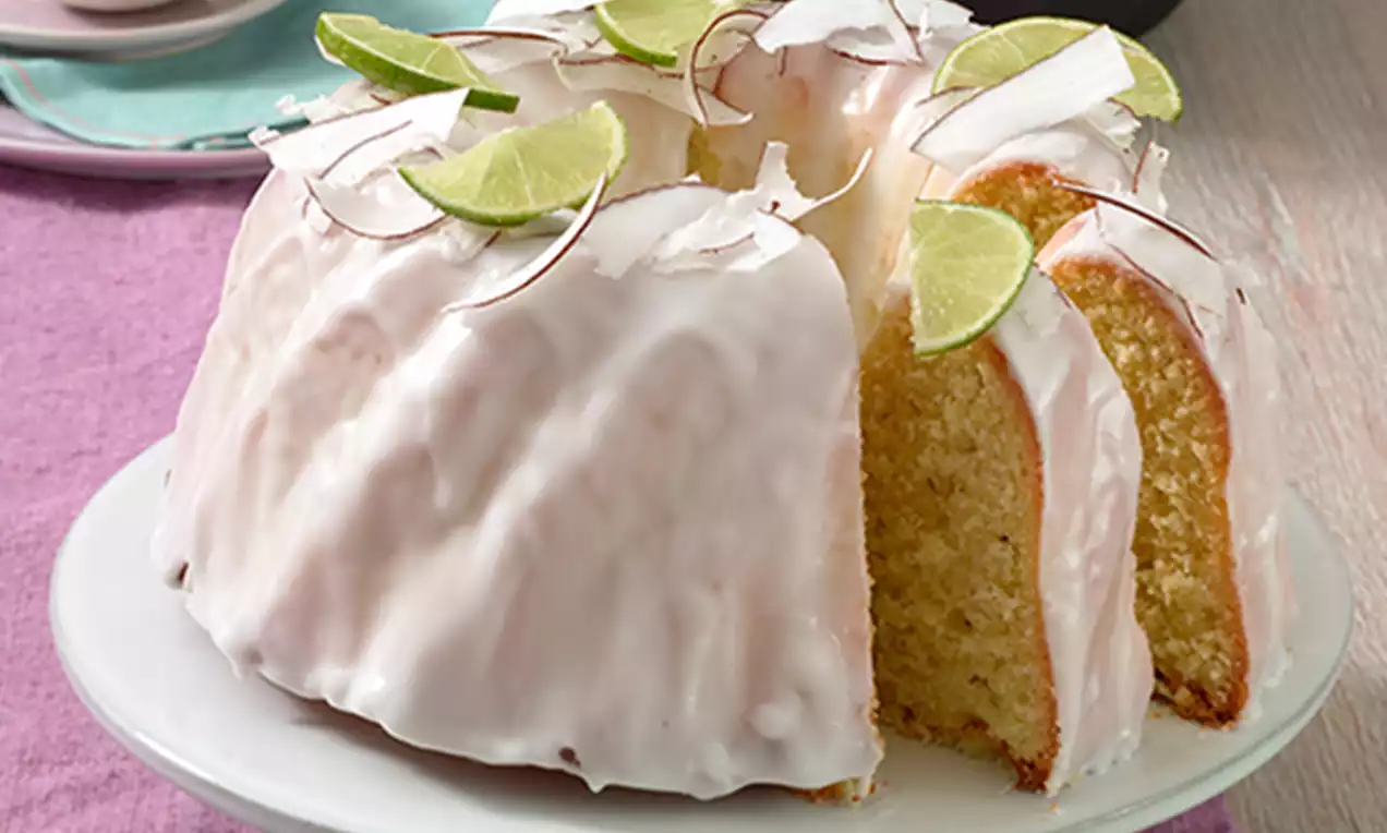 Luchtige tulbandcake met glanzende witte glazuurlaag, versierd met frisse limoenschijfjes en kokosvlokken op een wit taartplateau