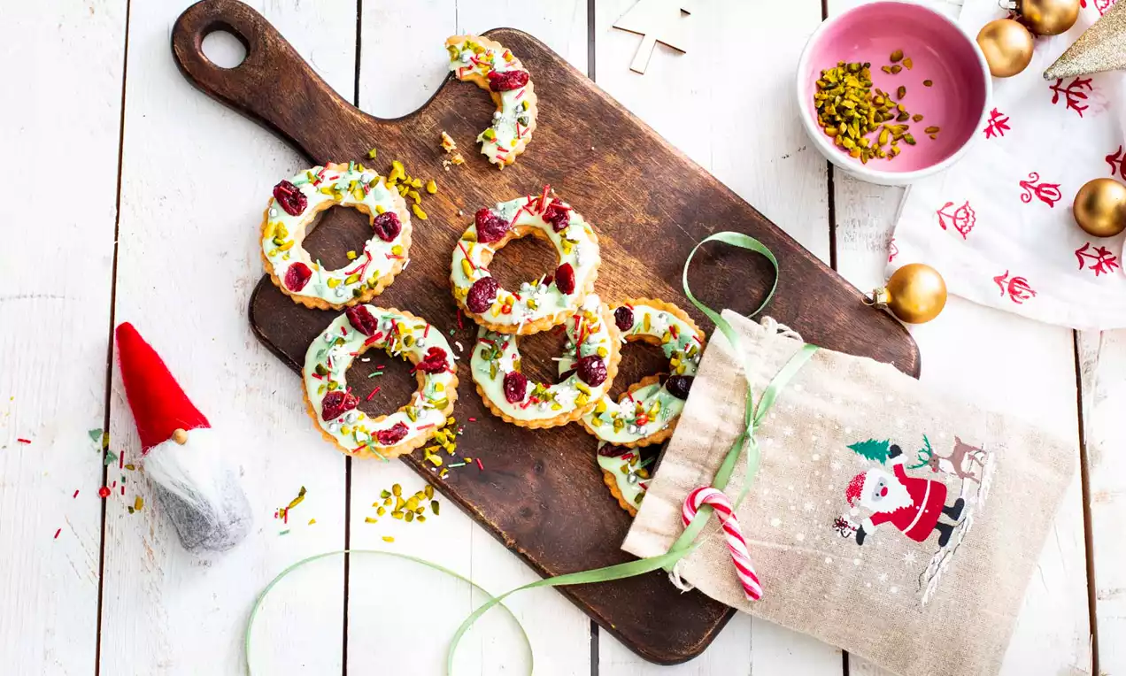 Zeltaini cepumu vainagi ar baltu glazūras kārti, rotāti sarkaniem augļiem un zaļām pistācijām, uz tumšas koka dēlīša blakus svētku maisiņam