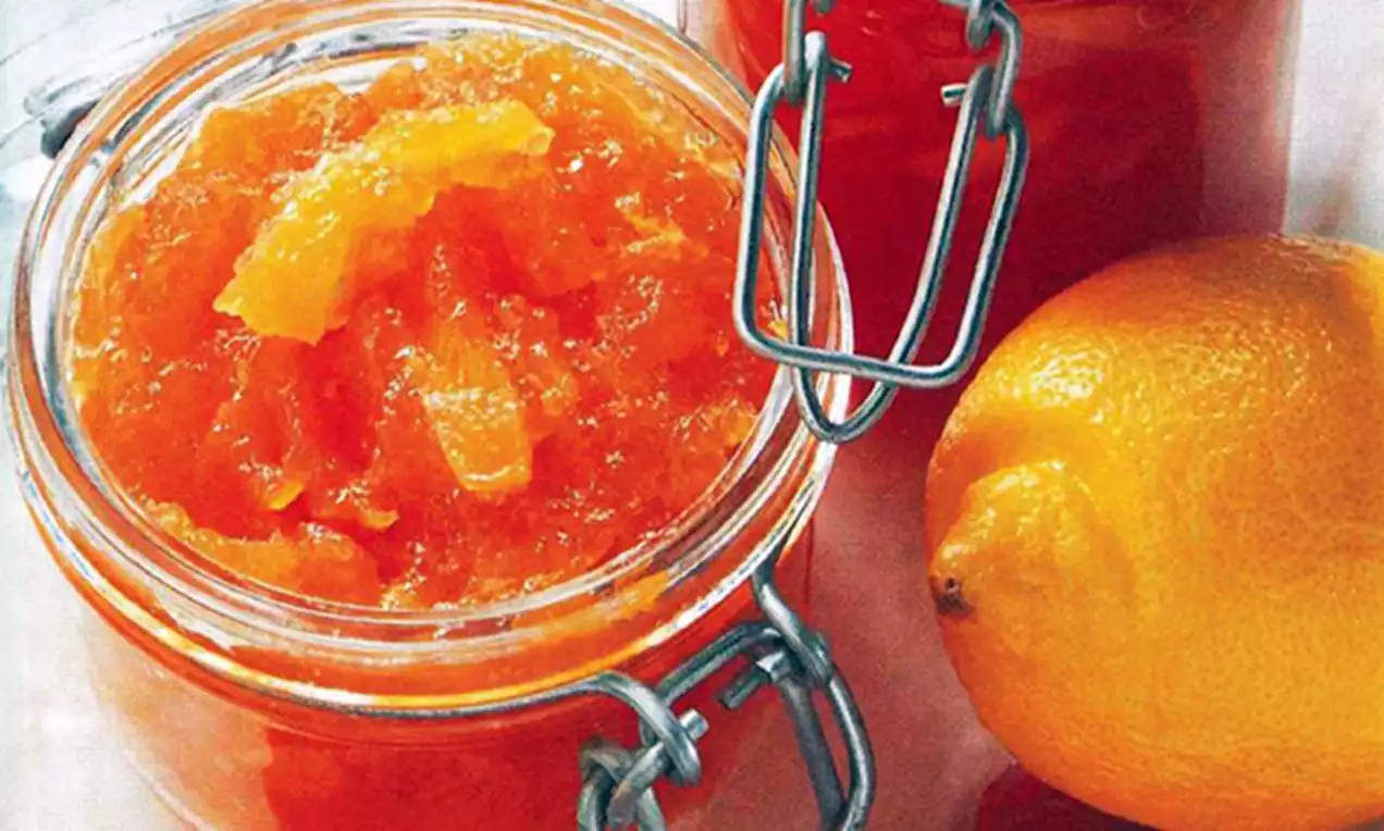 Een glazen pot gevuld met helder oranje abrikozenconfituur met stukjes fruit, naast een gele citroen en een rode pot met sluiting