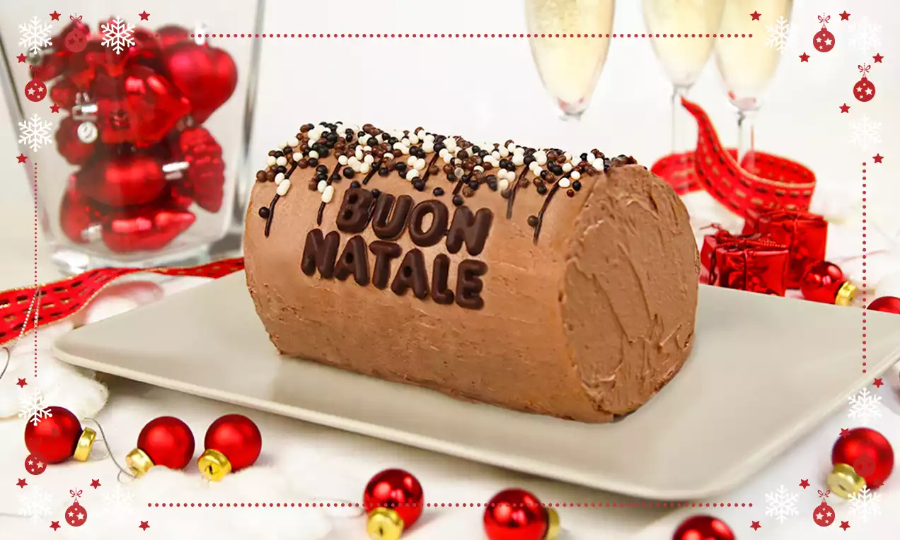 Tronchetto al cioccolato dalla superficie morbida e cremosa, decorato con scritta di cioccolato e granella, su un piatto bianco