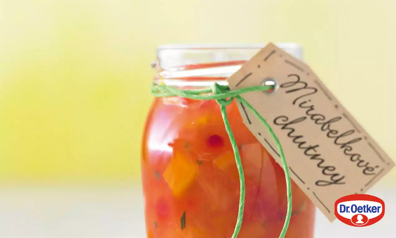 Sklenice s mirabelkovým chutney v odstínech oranžové a červené, zdobená zeleným provázkem a štítkem, na světlém pozadí