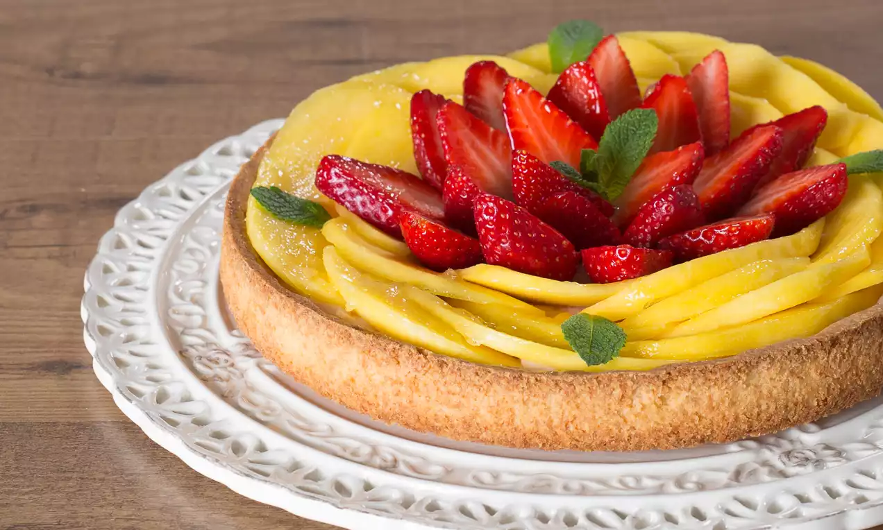 Crostata con base dorata friabile, decorata da fette di mango giallo intenso e fragole rosse su un piatto bianco intagliato