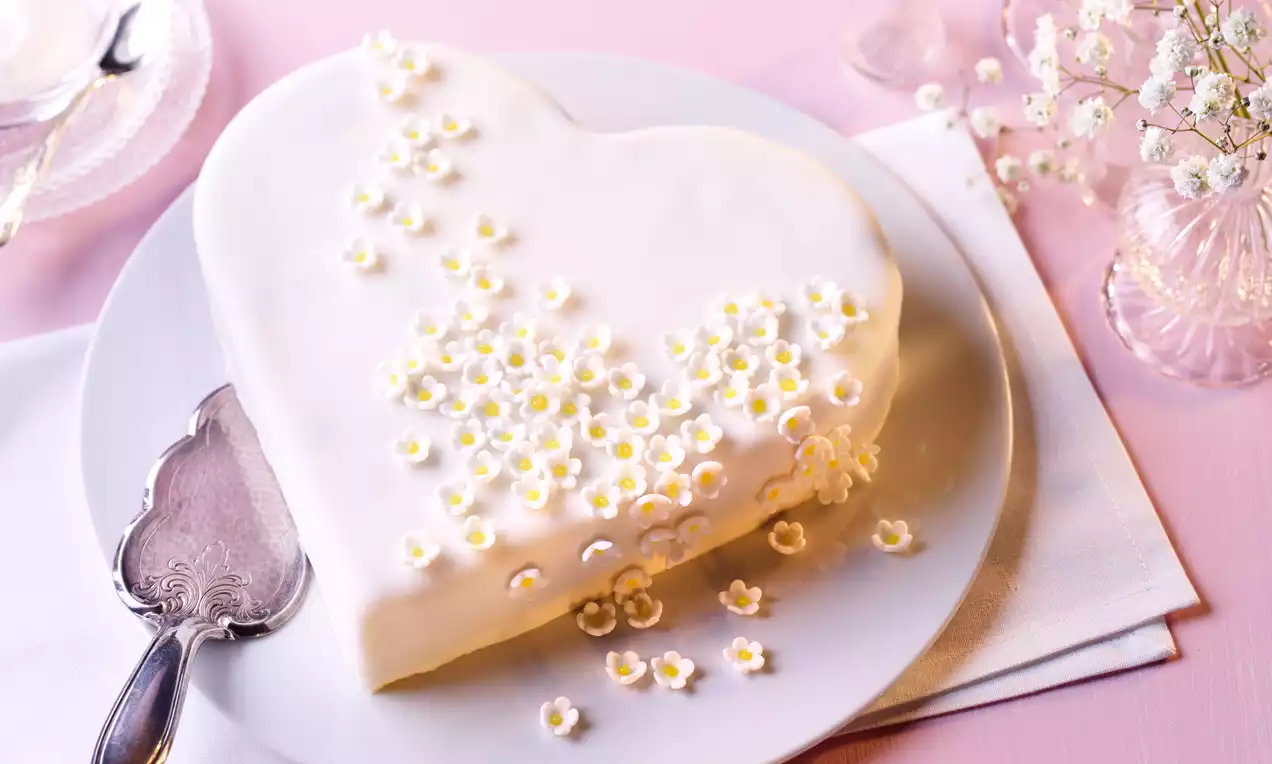 Herztorte mit weißer Glasur, verziert mit kleinen cremefarbenen Blüten, auf einem weißen Teller, neben Besteck und zarten Schleierkrautzweigen